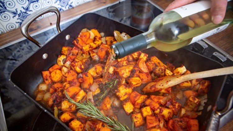 Eine Pfanne mit gewürfeltem Gemüse, darunter Süßkartoffeln, Zwiebeln und Rosmarin, wird mit Weißwein übergossen.