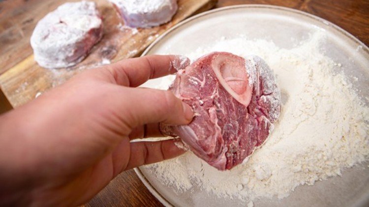 Eine Hand wendet ein Stück mehliertes Rindfleisch mit Knochen auf einem Teller voller Mehl, zwei weitere Stücke liegen im Hintergrund.