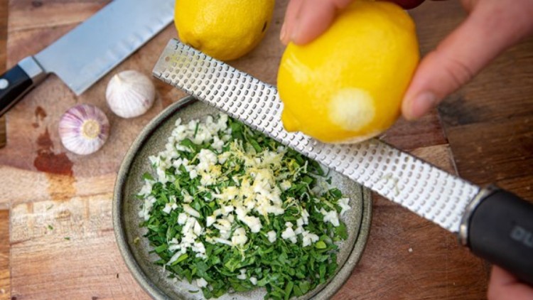 Eine Hand reibt Zitronenschale über eine Schale mit gehackter Petersilie, Knoblauch und Zwiebeln, daneben liegen Zitronen und Messer.