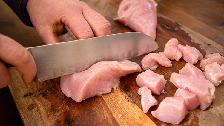 Eine Hand schneidet mit einem großen Messer rohes Hähnchenfleisch auf einem Holzbrett in Würfel.