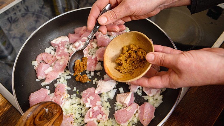 Eine Person gibt Gewürzpaste mit einem Löffel aus einer Schale zu rohem Fleisch und gehackten Zwiebeln in einer Pfanne.