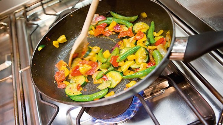 Bunte Gemüsestückchen wie Zucchini, Paprika und Zuckerschoten werden in einer schwarzen Pfanne mit Holzlöffel auf Gasherd gebraten.