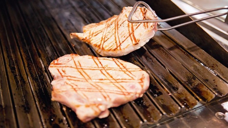 Zwei Schweinesteaks mit Grillstreifen liegen auf einem Rost, eines wird mit einer Grillzange gewendet.