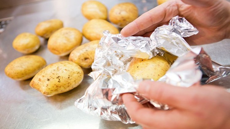 Eine Person wickelt eine rohe Kartoffel in Aluminiumfolie ein, weitere Kartoffeln liegen auf einer Metalloberfläche.