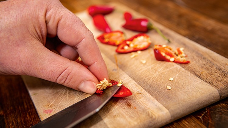 Eine Hand schneidet rote Chilischoten auf einem Holzschneidebrett, die Samen und Stücke der Schoten liegen daneben.