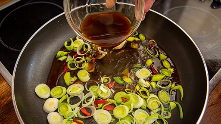 Eine Hand gießt braune Sauce aus einer Glasschale in eine Pfanne mit geschnittenem Lauch und roten Chili-Stückchen.