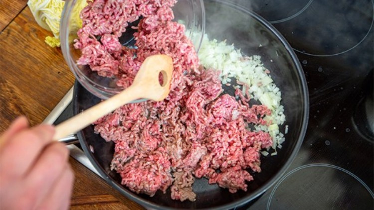 Rohes Hackfleisch und gehackte Zwiebeln werden in einer schwarzen Pfanne auf einem Herd mit einem Holzlöffel angebraten.