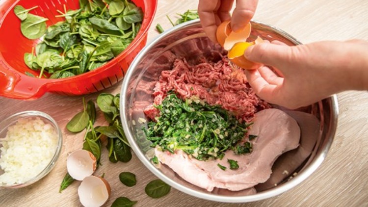 Eine Schüssel mit Hackfleisch, Spinat und Zwiebeln, daneben eine Hand, die ein Ei aufschlägt, auf einem Tisch mit weiteren Zutaten.
