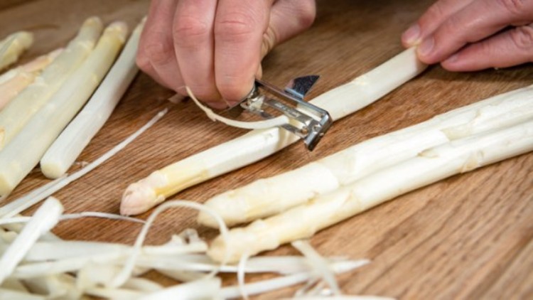 Hände schälen weißen Spargel auf einem Holzbrett mit einem Spargelschäler, Spargelschalen liegen daneben.