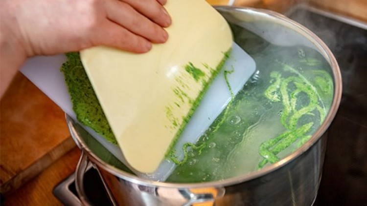 Gehackter, grüner Spinat wird von einem Schneidebrett in einen glänzenden Kochtopf mit kochendem Wasser gegeben.