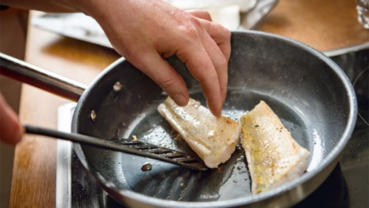 Eine Hand wendet ein goldbraun gebratenes Fischfilet in einer schwarzen Pfanne, daneben liegt ein weiteres Fischfilet.