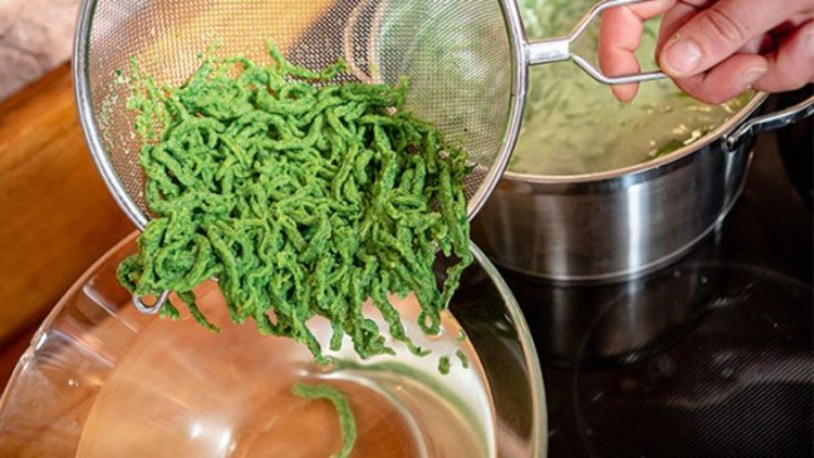 Grüner Spätzleteig wird in einem Sieb über einer Glasschüssel abgegossen, im Hintergrund steht ein Topf mit heißem Wasser.