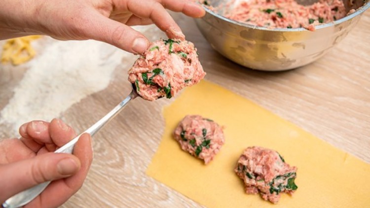 Eine Hand formt mit einem Löffel eine Füllung aus Hackfleisch und Kräutern, die auf gelben Nudelteig gelegt wird.