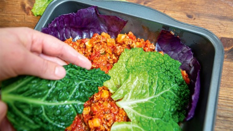Eine Hand legt ein großes grünes Wirsingblatt auf eine Schicht aus Hackfleisch-Tomatensoße in einer dunkelgrauen Backform.