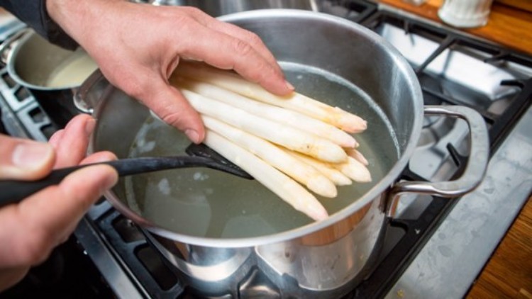 Eine Hand hält weißen Spargel über einen Kochtopf mit heißem Wasser, daneben ist eine schwarze Kelle zu sehen.