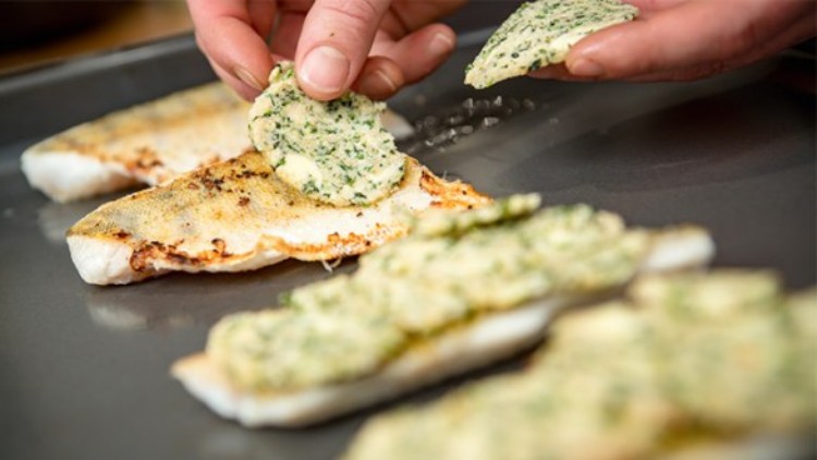 Gegrilltes Fischfilet auf Backblech wird mit Kräuterbutterscheiben von einer Hand belegt.