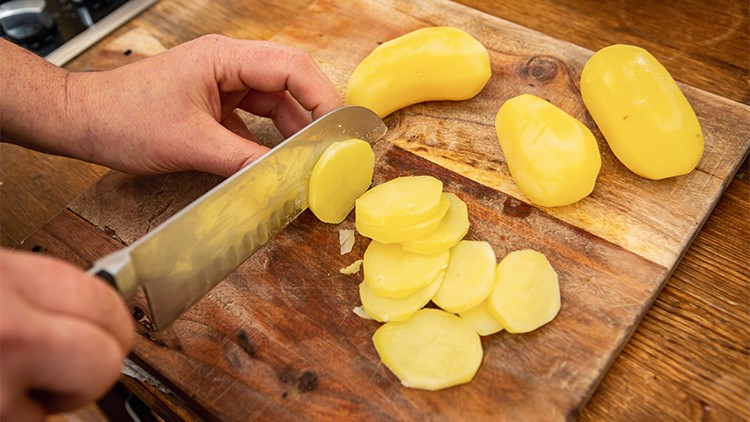Eine Hand schneidet geschälte gelbe Kartoffeln mit einem Messer in dünne Scheiben auf einem Holzbrett.