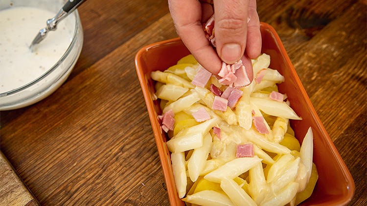 Hand streut fein geschnittenen Schinken auf Kartoffeln, Spargel und Käse in einer orangefarbenen Auflaufform.