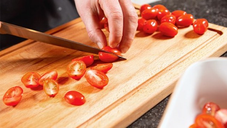 Eine Hand schneidet mit einem Messer rote Cherrytomaten auf einem Holzbrett, weitere Tomaten liegen daneben.