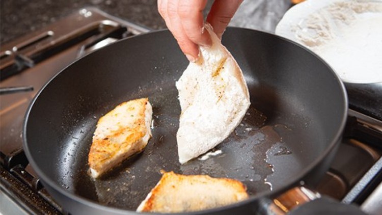 Eine Hand legt ein bemehltes Fischfilet in eine schwarze Pfanne, in der bereits zwei gebratene Stücke liegen.