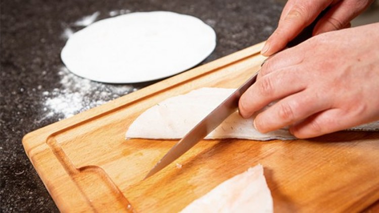 Hände schneiden mit einem Messer ein Fischfilet auf einem Holzbrett, im Hintergrund liegt ein runder Teigfladen.