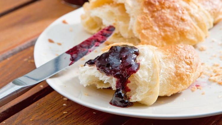 Auf einem weißen Teller liegt ein aufgeschnittenes Croissant mit roter Marmelade, daneben ein Messer und ein Marmeladenglas.
