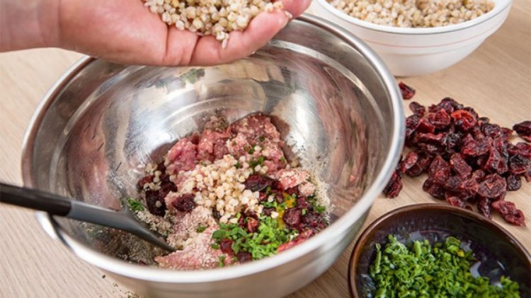 Hand fügt Hirse zu Hackfleisch, Kräutern und getrockneten Cranberrys in eine Metallschüssel, daneben Zutaten in kleinen Schalen.