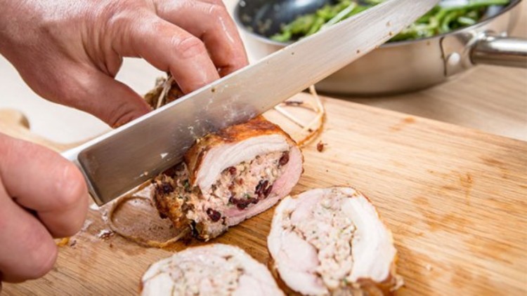 Person schneidet gefülltes Fleisch mit Messer auf Holzbrett, dahinter steht eine Pfanne mit grünem Gemüse.