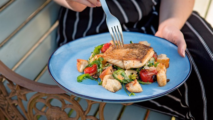 Gebratenes Fischfilet auf Salat mit Croutons, Kirschtomaten und Spinat auf einem blauen Teller, Hand mit Gabel hält ein Stück.