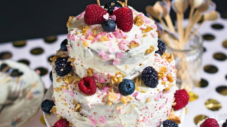 Zweistöckige Torte mit weißer Creme, Beeren, Nüssen, rosa Zuckerherzen und zwei goldenen Herz-Dekorationen auf der Spitze.