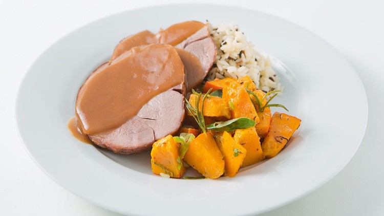 Auf einem weißen Teller liegen zwei Scheiben Fleisch mit Bratensoße, gebackener Kürbis und eine Portion Wildreis.