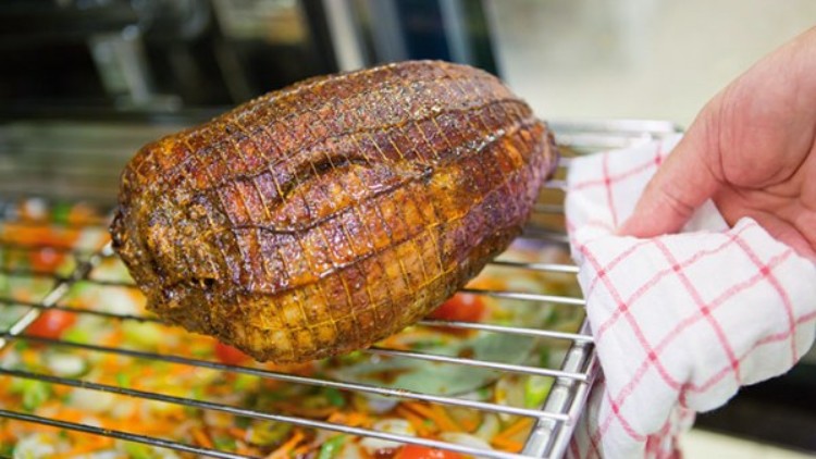 Ein Braten in Gewürzkruste liegt auf einem Ofengitter, darunter Gemüse, eine Hand hält ein rot-weiß kariertes Tuch am Gitter.