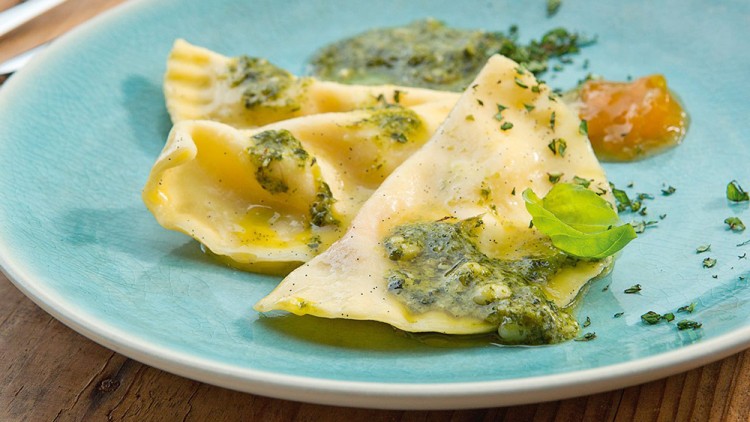 Drei gefüllte Teigtaschen mit grünem Pesto, Kräutern und einem Klecks gelblicher Soße auf einem türkisfarbenen Teller.