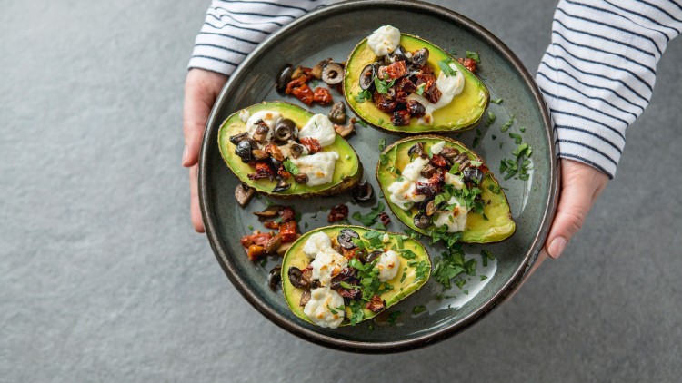 Gefüllte Avocadohälften mit Käse, Oliven, getrockneten Tomaten und Kräutern auf einem grauen Teller in Händen mit gestreiftem Pullover.