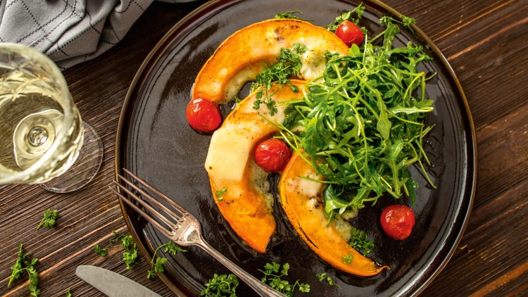Gegrillte Kürbisspalten mit geschmolzenem Käse, Kirschtomaten und frischem Rucola auf einem dunklen Teller, daneben ein Weinglas.