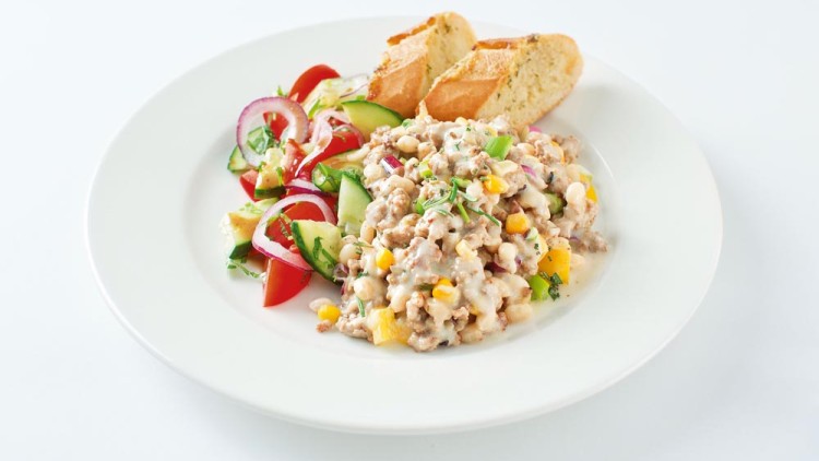 Ein Teller mit Hackfleischpfanne, Mais und Gemüse in heller Soße, dazu Tomatensalat mit Gurke und Baguettescheiben.