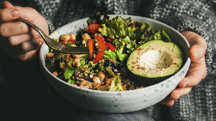 Eine Person hält eine Schale mit Salat, Kichererbsen, Quinoa, Tomaten, Blattsalat und einer halbierten Avocado mit Würzung.