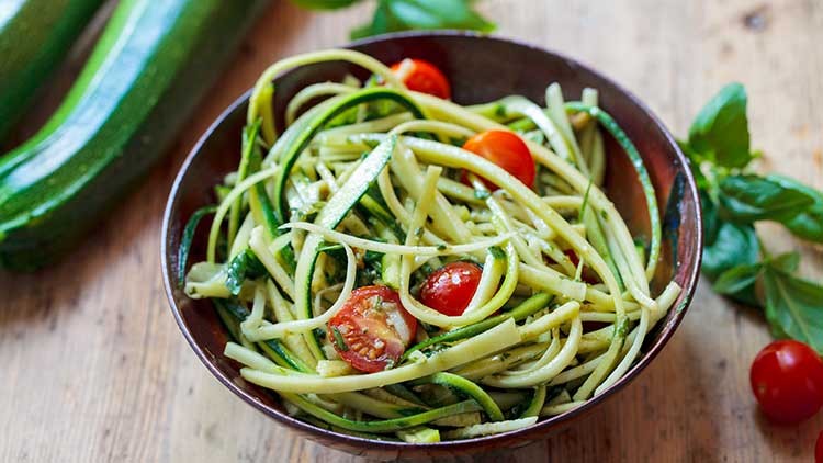 Eine Schale mit Zucchininudeln, halbierten Kirschtomaten und Basilikumblättern auf einem Holztisch.