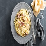 Spaghetti mit heller Soße, geriebenem Käse und Schinken auf einem grauen Teller, daneben Brot auf hellem Stoff.