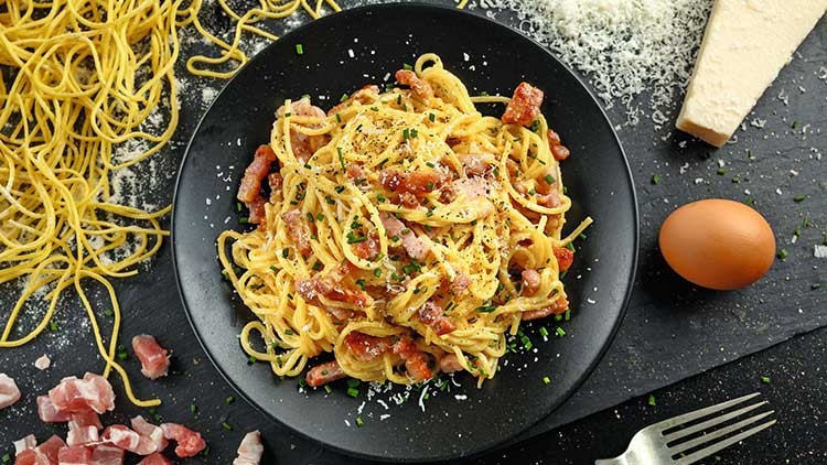 Ein schwarzer Teller mit Spaghetti, Speck, geriebenem Käse und Schnittlauch, daneben Parmesan, ein Ei und rohe Nudeln.
