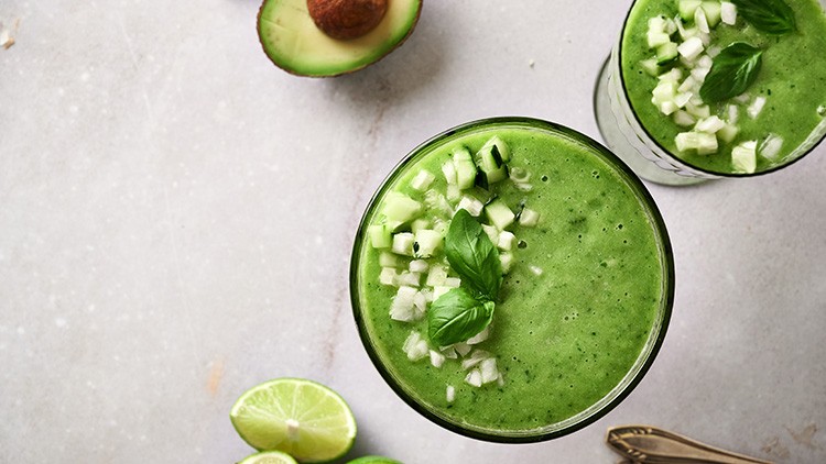 Zwei Gläser grüner Smoothie mit Gurkenwürfeln, Basilikumblättern, daneben Avocadohälfte und Limettenscheiben auf hellem Untergrund.