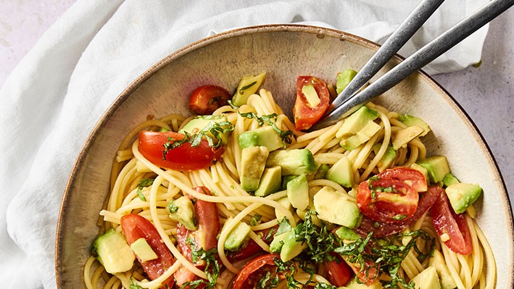 Eine Schale mit Spaghetti, Avocado-Würfeln, Tomatenstücken und gehackter frischer Kräuter, mit zwei Besteckteilen darauf.