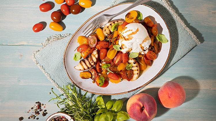 Ein Teller mit Tomatensalat, gegrilltem Brot, Basilikum, Burrata und Balsamico, daneben Pfirsiche, Kräuter und Gewürze.