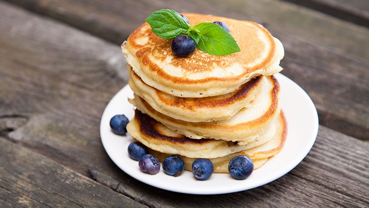 Ein Stapel Pfannkuchen mit Blaubeeren und einem Minzblatt auf einem weißen Teller, auf Holztisch liegend.