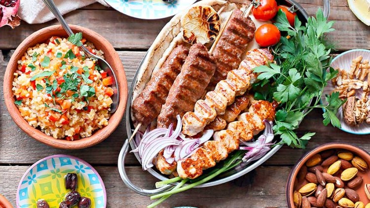 Grillgemüse, Reisgericht, Fleischspieße und Dips auf Holztisch mit frischem Gemüse, Nüssen, Kräutern und Fladenbrot.