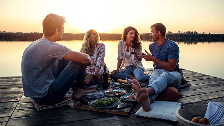 Vier Personen sitzen auf einem Steg am Wasser bei Sonnenuntergang, vor ihnen ein Picknick mit Obst, Brot und Getränken.
