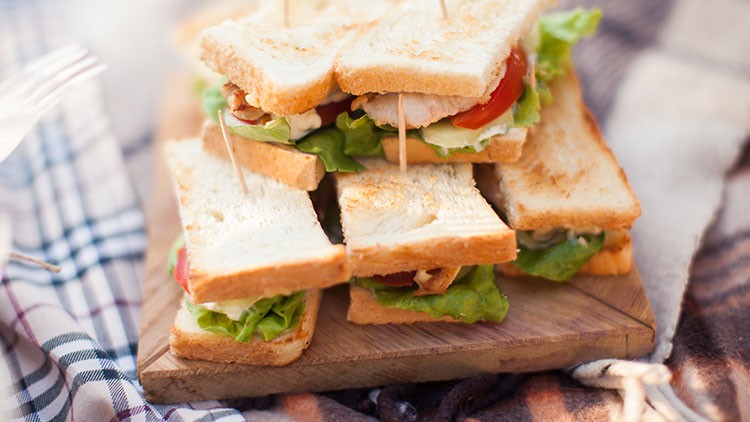 Mehrere Sandwich-Viertel mit Toastbrot, Salat, Tomaten und Holzspießen liegen auf einem Holzbrett, daneben liegt ein kariertes Tuch.