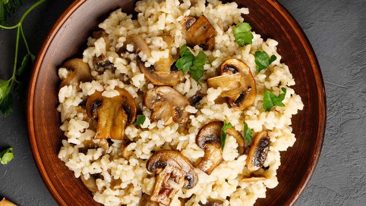 Eine braune Schale mit cremigem Pilzrisotto, dekoriert mit Petersilienblättern, steht auf einer grauen Steinoberfläche.