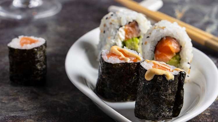 Mehrere Sushirollen mit Lachs, Reis, Avocado und Nori auf weißem Teller, daneben Holzstäbchen auf dunklem Untergrund.