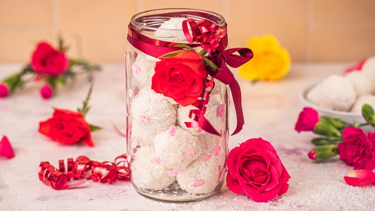 Ein Glas mit weißen Kokosbällchen, verziert mit rotem Band und einer roten Rose, umgeben von Blumen und Kokosflocken.
